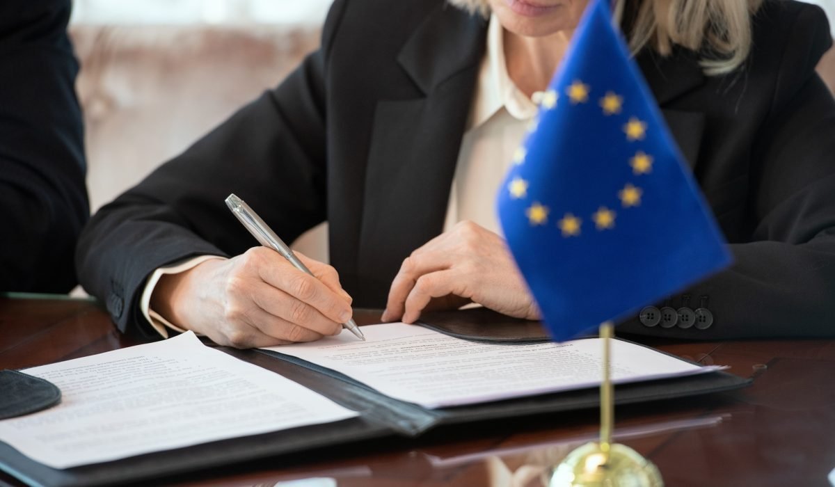 Flag of European Union on table against mature female delegate signing contract after negotiating and making deal by table in boardroom