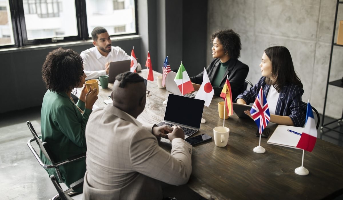 International Business Meeting. Multicultural Coworkers, Businessmen And Businesswomen, Sitting At Table With Flags Of Different Countries In Modern Office Indoor. Corporate Diversity Concept