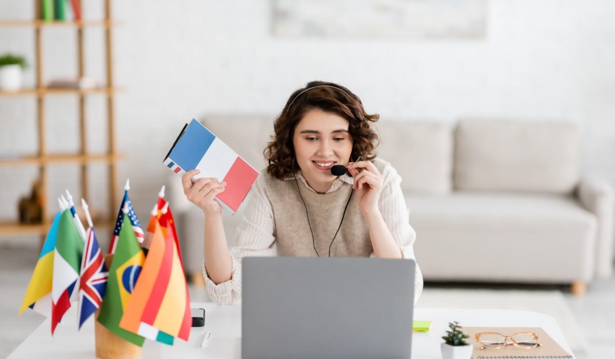 happy language teacher in headset holding French textbook near international flags during online lesson on laptop